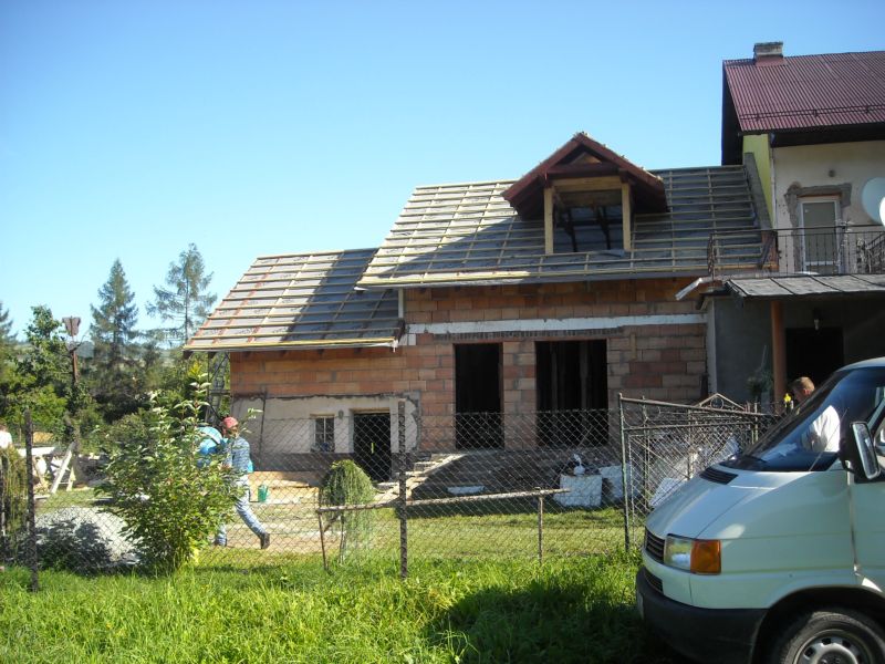 Budowa domu z czerwonej cegły w trakcie realizacji, widoczne przygotowania do położenia dachu, na pierwszym planie ogrodzenie i trawa, w tle częściowo widoczny samochód dostawczy.